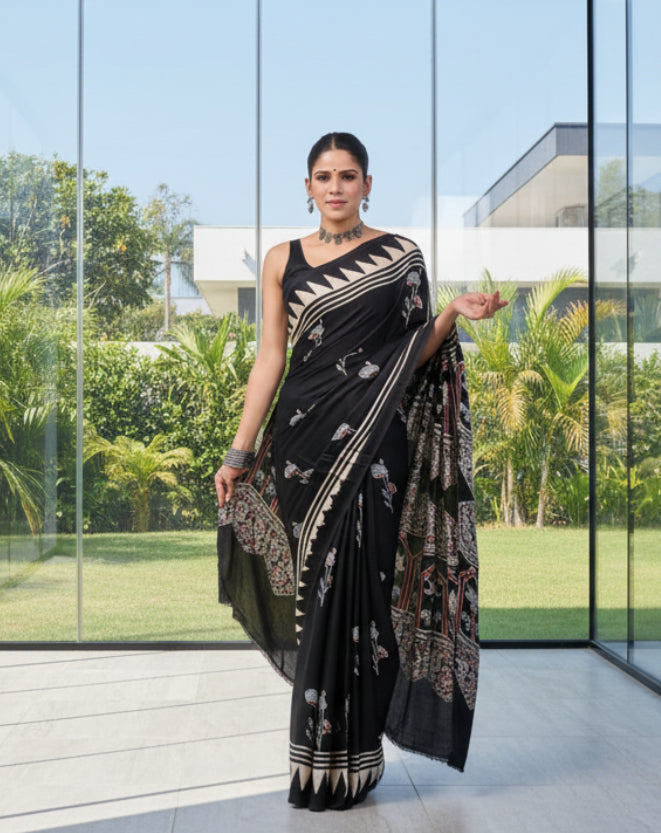 Woman in a black and white patterned saree standing in front of a glass wall with greenery outside.